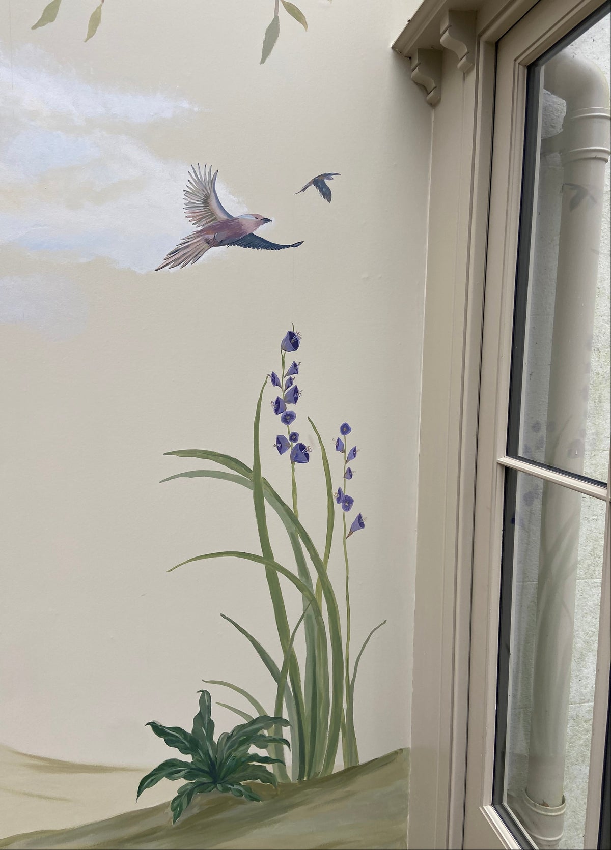 A hand-painted mural on a cream-colored wall depicting green reeds with purple flowers, a flying bird, and a small bird in flight, positioned next to a window frame.