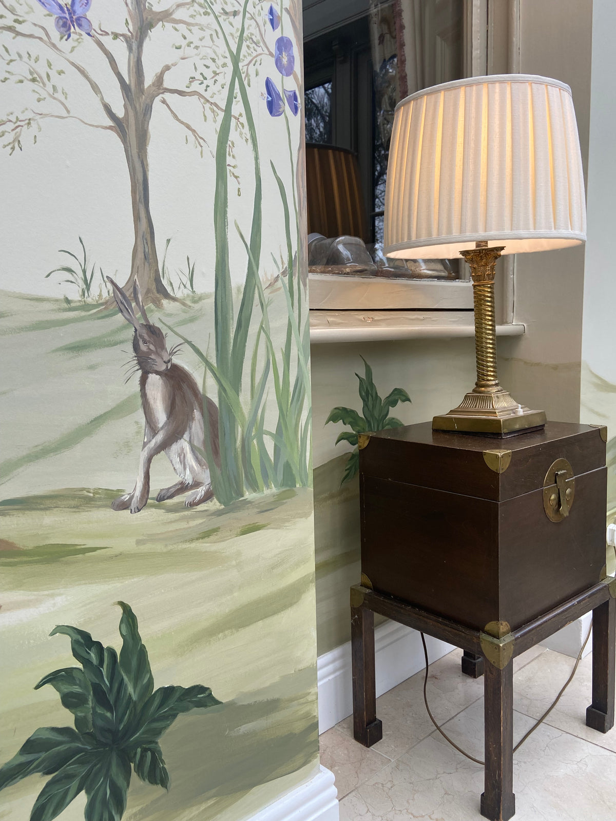 A dark brown wooden side table with brass corner brackets and a pleated white lampshade, styled in a room next to a hand-painted mural of a hare in a grassy landscape.