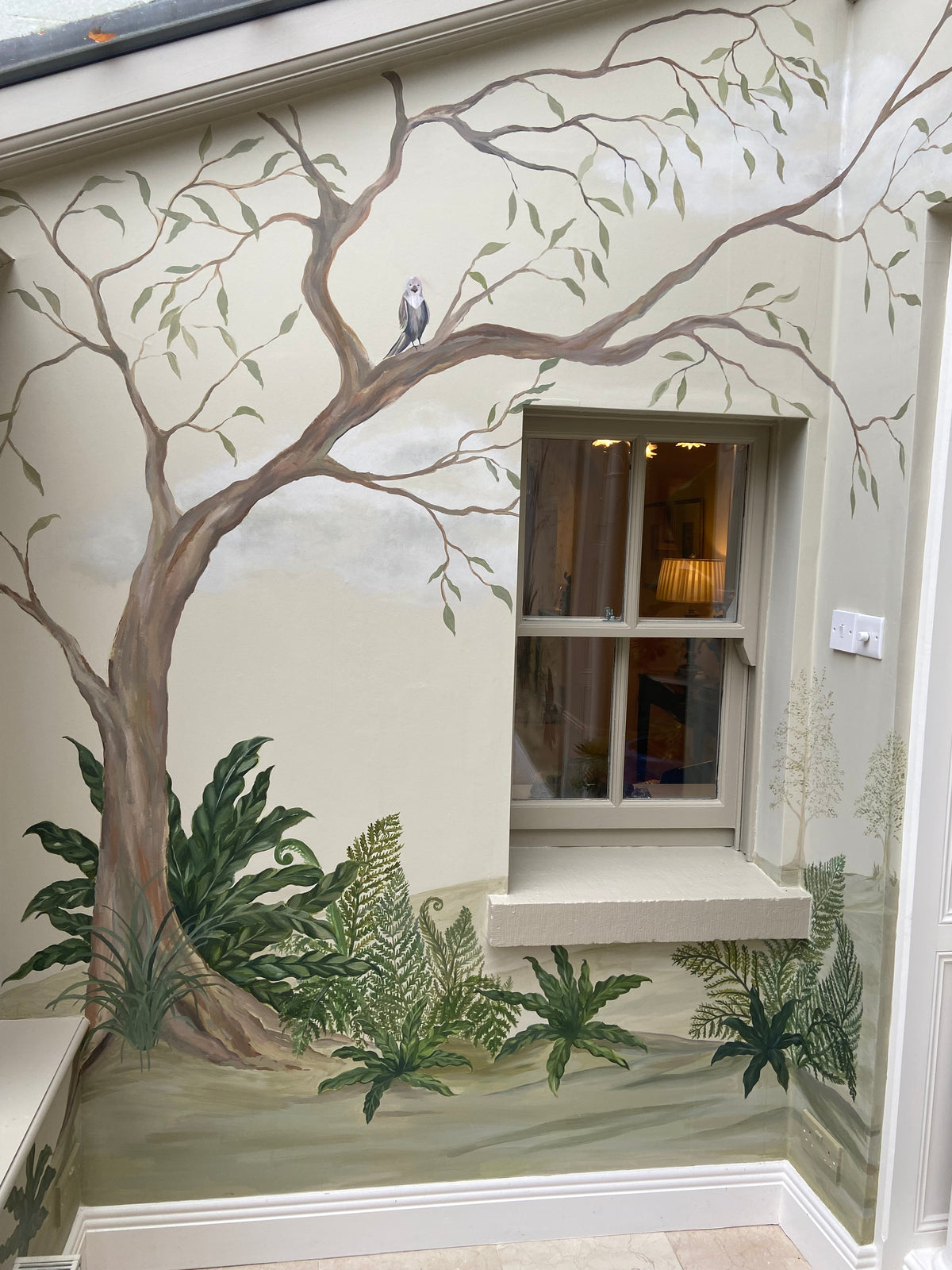 A large hand-painted nature mural on a wall, featuring a large tree with a small bird perched on its branch, green ferns, and a window set into the painted scene.