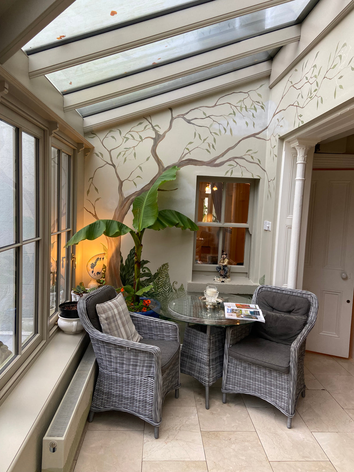 A furnished sunroom corner with two gray wicker armchairs and a round glass-top side table, set against a wall with painted tree branches and a large potted banana plant. A skylight and windows illuminate the space.