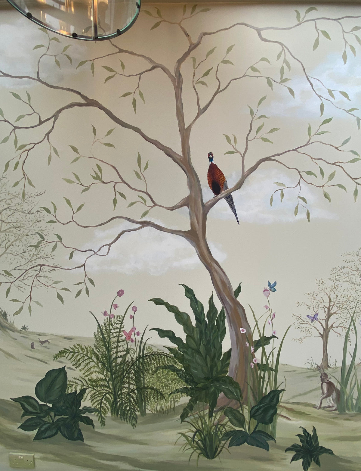 A large hand-painted mural on a wall depicting a forest scene with a central tree, a pheasant bird, green foliage, flowers, and a deer, shown in an interior room setting.