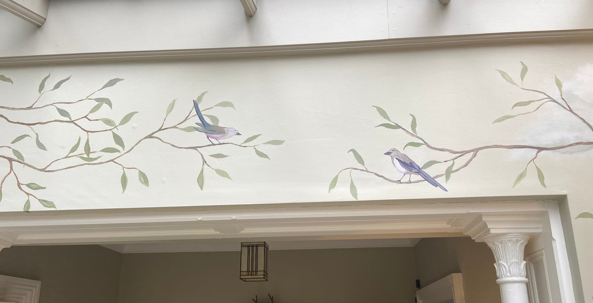 A hand-painted mural of two bluebirds perched on leafy branches, set against a warm cream-colored wall above a doorway.