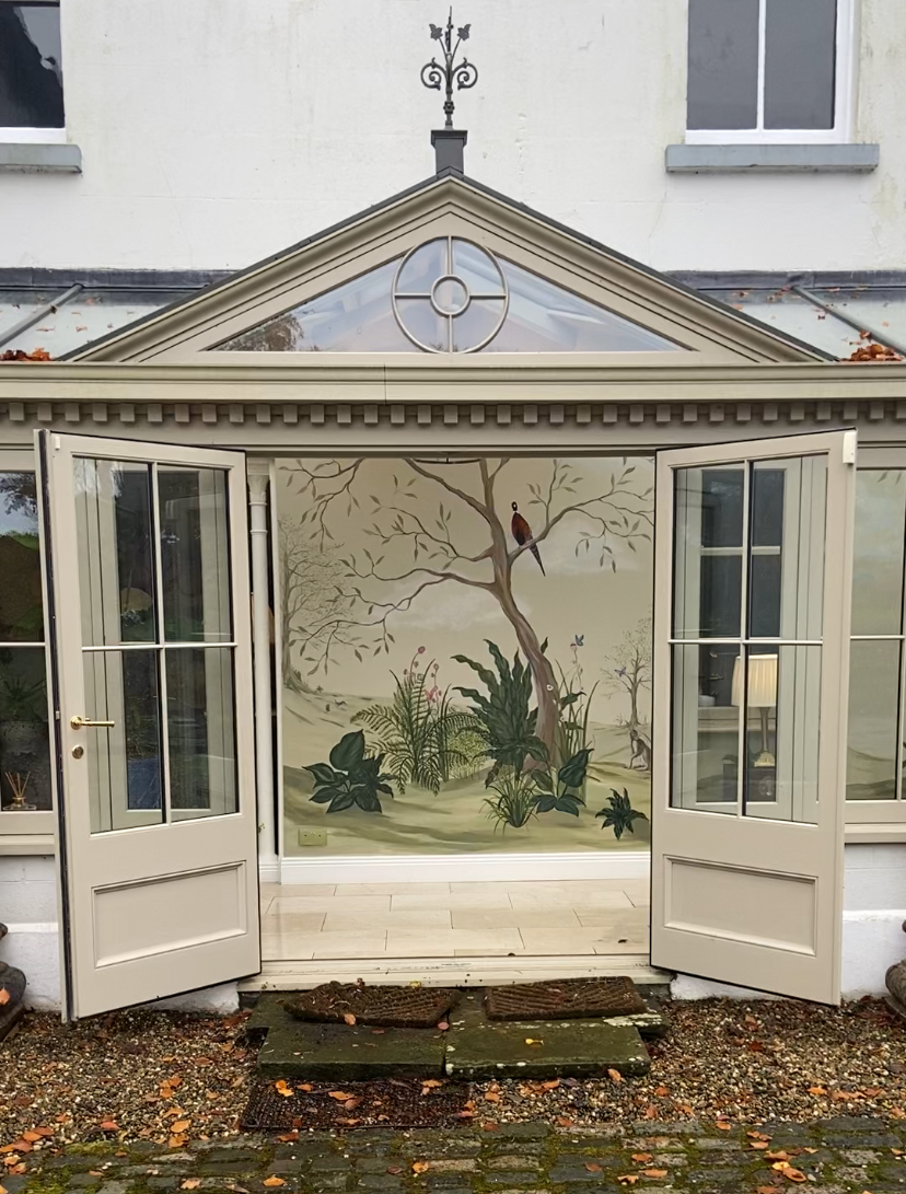 A large cream-colored conservatory extension with open French doors, featuring a hand-painted nature mural of a tree and birds on the back wall, set against a white building facade with gravel and fallen leaves in the foreground.